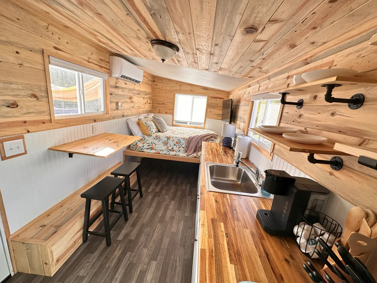 A warm, open-concept interior featuring a cozy bed, a small dining table, a kitchen counter, and generous windows that flood the space with natural light. The layout is inviting, with everything within reach for both relaxation and social interaction. The wood elements and soft textiles enhance the comfort, while the windows offer scenic glimpses of the outdoors.