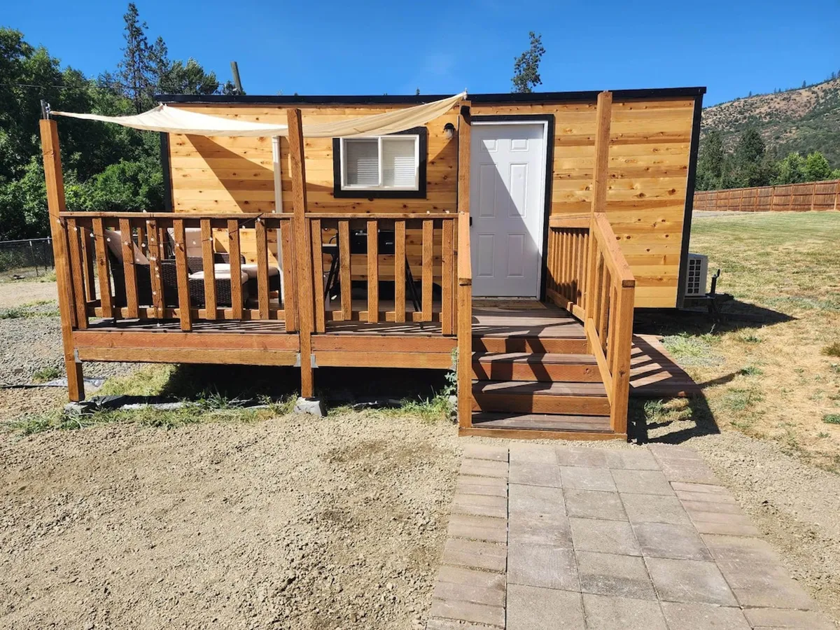 This image captures the exterior of the cabin, focusing on a welcoming wooden deck with stairs leading up to the door. The space is adorned for relaxation, with seating options perfect for enjoying surrounding nature, and the entryway is visible as a warm transition point between inside and out. The scene is set during golden hour, offering a cozy and inviting atmosphere.