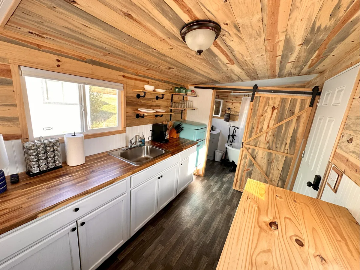 A warmly lit compact kitchen featuring a wooden countertop, deep farmhouse sink, sliding barn door, and a retro green fridge. The scene suggests a cozy and functional layout perfect for preparing meals and gathering in an intimate space.