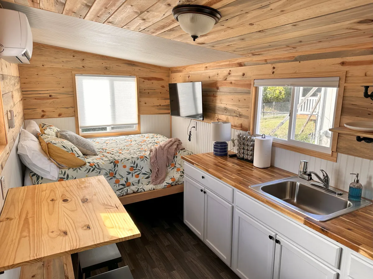 The image showcases a cozy interior of a tiny house, featuring a sunlit bed with a floral comforter, a compact wooden kitchen counter with a sink, and a TV mounted on the wall. The space is thoughtfully designed for comfort and relaxation, inviting guests to unwind, cook, and enjoy peaceful moments.
