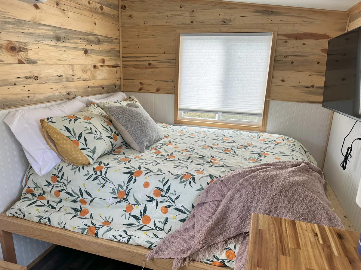 A bright, inviting nook with a comfortable bed dressed in floral bedding, rich wooden wall panels, and a large window letting in streams of daylight. The window frames a serene view outside, enhancing the room's tranquil, lived-in ambiance. A TV mounted on the wall and thoughtful touches like reading lamps make the space ideal for relaxation and leisure.